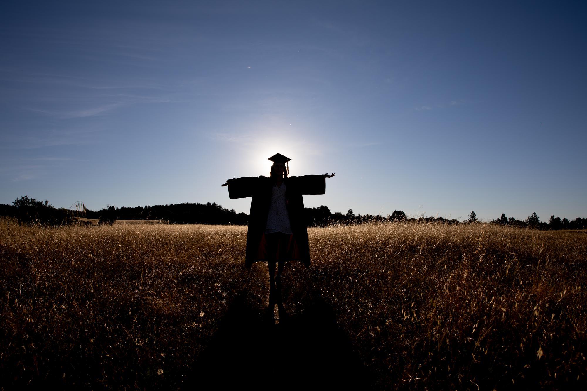 Person standing in a field with the sun behind them, wearing their cap and gown.